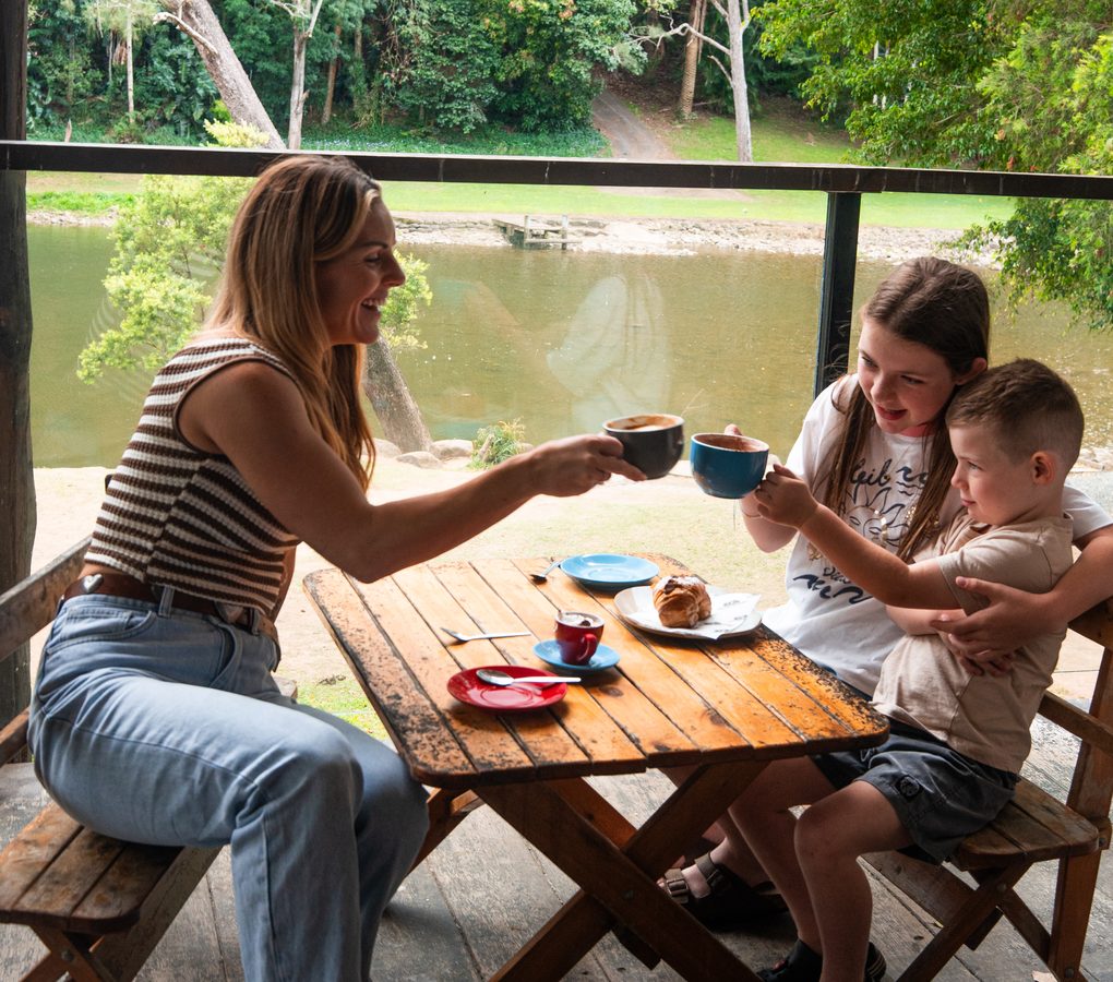 A family clinking cups of coffee and hot chocolate at a table on the Rivermill Café deck, the river behind them