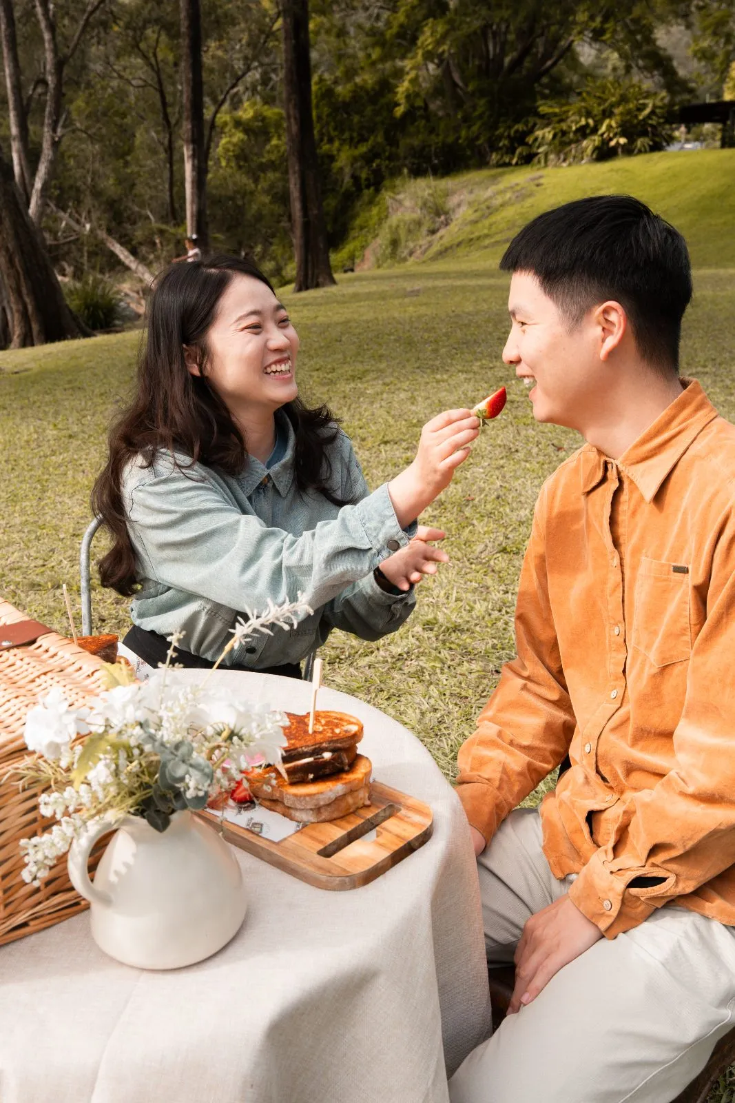 A couple laughing over a picnic on the lawns at Historic Rivermill, sharing strawberries from a hamper
