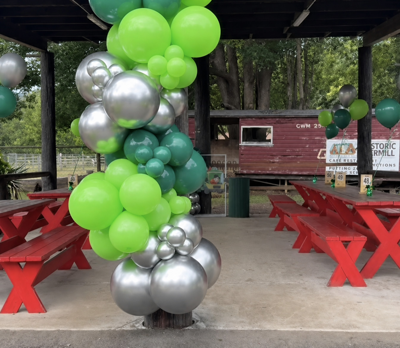 The Red Tables set for a birthday party, with a green and silver balloon arch under the undercover picnic area