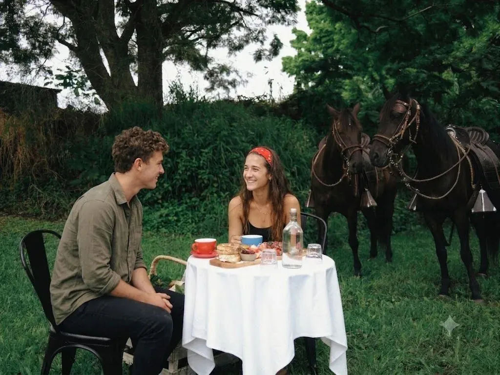A couple sharing food and drinks at a small table on the grass, with two saddled horses standing behind them at Historic Rivermill