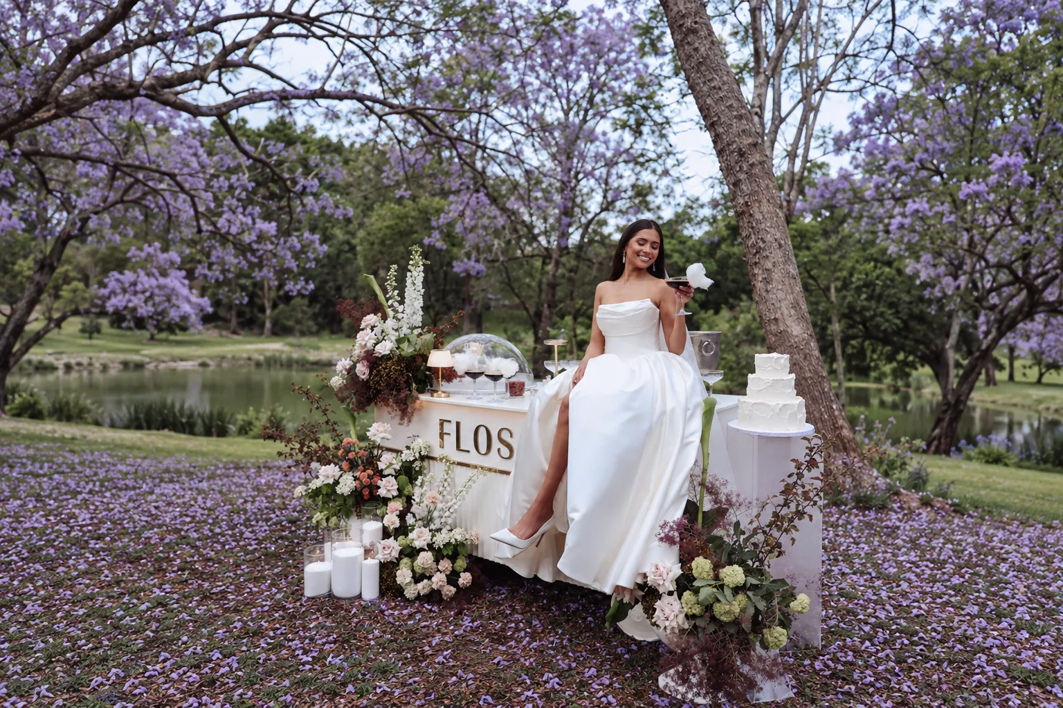 A bride seated at a floral drinks bar with a tiered cake, jacaranda petals carpeting the grass and the river behind, at Historic Rivermill