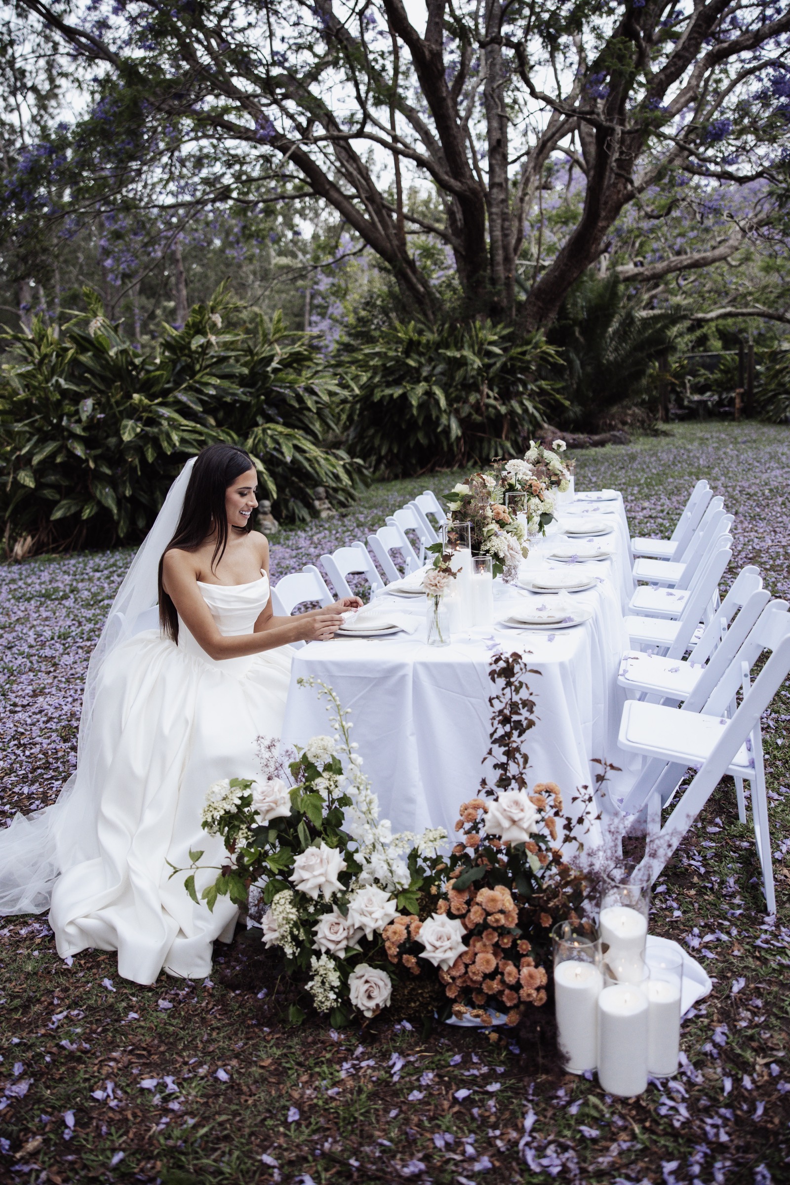A bride at a long reception table set under a flowering jacaranda tree, purple petals across the lawn at Historic Rivermill