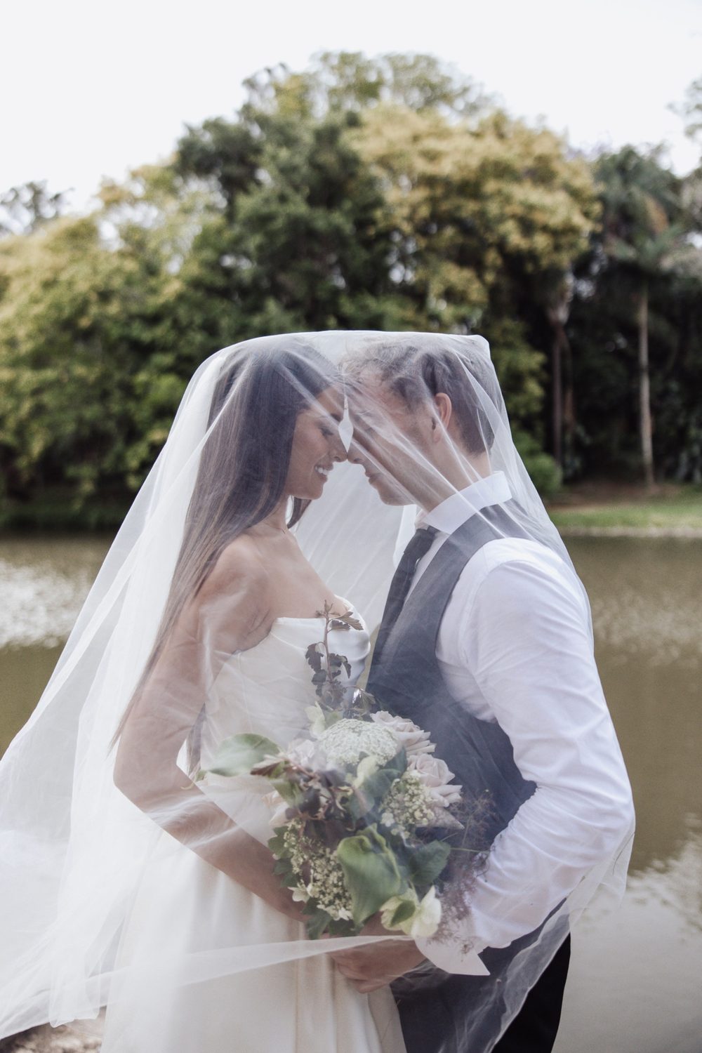 Bride and groom nose to nose beneath her veil by the water